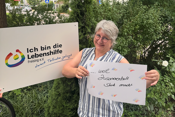 Petra Neumaier feiert die Demokratie, weil Zusammenhalt stark macht. Foto: Lebenshilfe Eine Frau mit Brille steht im Grünen und hält ein Schild, daneben hält jemand ein Schild "Ich bin die Lebenshilfe Freising e.V.