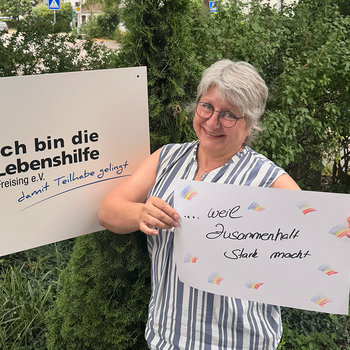 Petra Neumaier feiert die Demokratie, weil Zusammenhalt stark macht. Foto: Lebenshilfe Eine Frau mit Brille steht im Grünen und hält ein Schild, daneben hält jemand ein Schild "Ich bin die Lebenshilfe Freising e.V.