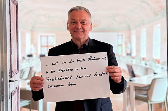 Franz Heilmeier, stellvertretender Landrat, feiert die Demokratie weil sie der beste Rahmen ist, in dem Menschen in ihrer Verschiedenheit fair und friedlich zusammen leben. Foto: Kreisjugendring Ein Mann im schwarzen Sakko steht in der Tür zu einem hellen Sitzungssaal und hält lächelnd ein Schild.