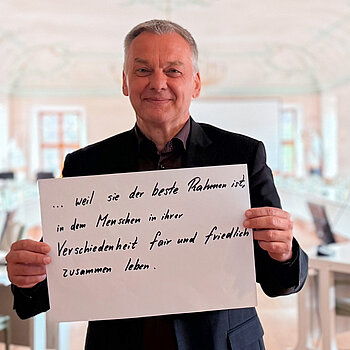 Franz Heilmeier, stellvertretender Landrat, feiert die Demokratie weil sie der beste Rahmen ist, in dem Menschen in ihrer Verschiedenheit fair und friedlich zusammen leben. Foto: Kreisjugendring Ein Mann im schwarzen Sakko steht in der Tür zu einem hellen Sitzungssaal und hält lächelnd ein Schild.