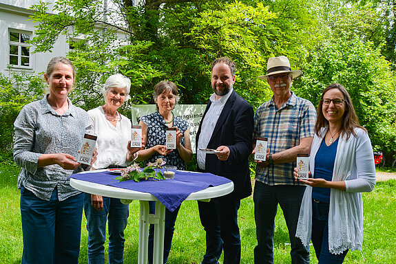 zwei Männer und vier Frauen stehen nebeneinander und zeigen die Schokoladentafeln. Davor ein Stehtisch, dahinter grüne Büsche und ein Gebäude 