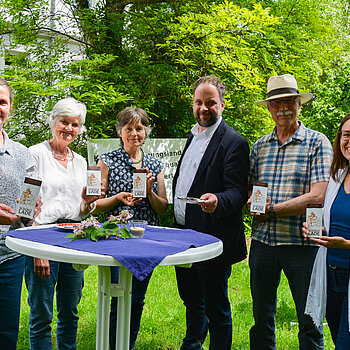 zwei Männer und vier Frauen stehen nebeneinander und zeigen die Schokoladentafeln. Davor ein Stehtisch, dahinter grüne Büsche und ein Gebäude 