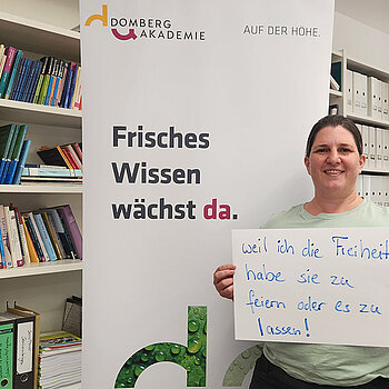Kathrin Steger-Bordon (Domberg Akademie) feiert, weil sie die Freiheit hat zu feiern oder es zu lassen. Foto: Domberg Akademie Eine Frau mit dunklen Haaren steht vor einem Bücherregal und neben einem Rollup mit einem Schild in der Hand.