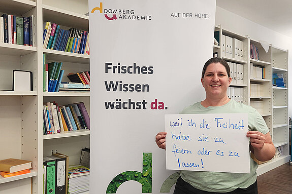 Kathrin Steger-Bordon (Domberg Akademie) feiert, weil sie die Freiheit hat zu feiern oder es zu lassen. Foto: Domberg Akademie Eine Frau mit dunklen Haaren steht vor einem Bücherregal und neben einem Rollup mit einem Schild in der Hand.
