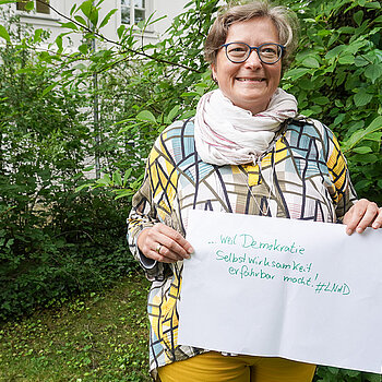 Christine Albrecht hält ein Schild mit der Aufschrift: "weil Demokratie Selbstwirksamkeit erfahrbar macht."