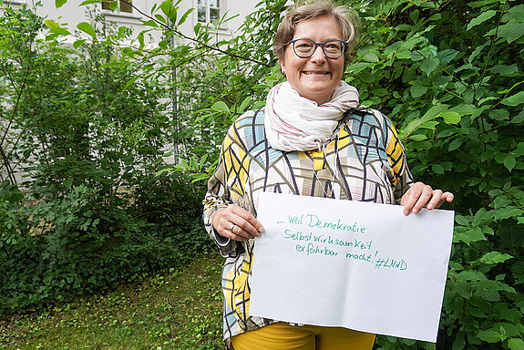 Christine Albrecht hält ein Schild mit der Aufschrift: "weil Demokratie Selbstwirksamkeit erfahrbar macht."