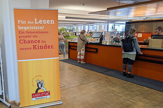 Mentor Leselernpaten warben in der Stadtbibliothek. Foto: Stadt Freising ein großes oranges Rollup "Für das Lesen begeistern!" steht vor dem Ausleihschalter der Bibliothek.
