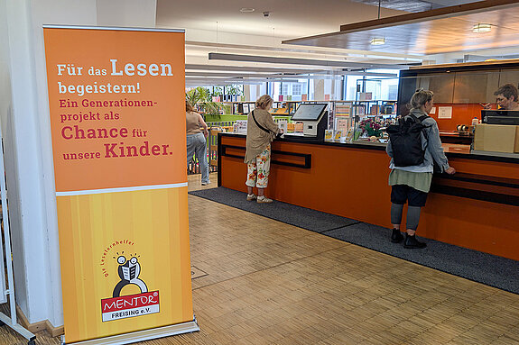 Mentor Leselernpaten warben in der Stadtbibliothek. Foto: Stadt Freising ein großes oranges Rollup "Für das Lesen begeistern!" steht vor dem Ausleihschalter der Bibliothek.