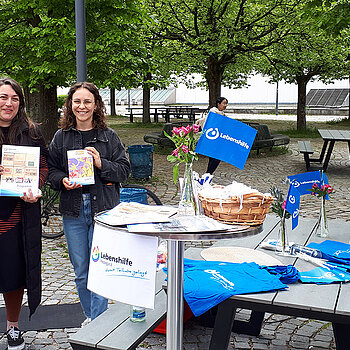 2 Frauen halten Broschüren hoch und stehen neben einem Tisch mit vielfältigem Material