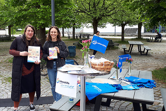 2 Frauen halten Broschüren hoch und stehen neben einem Tisch mit vielfältigem Material