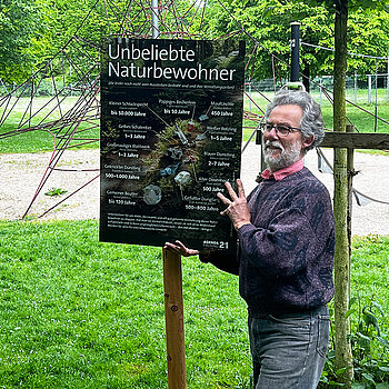 Ein Mann steht in einer Grünanlage und hält lächelnd ein großes Schild an einen Pfahl