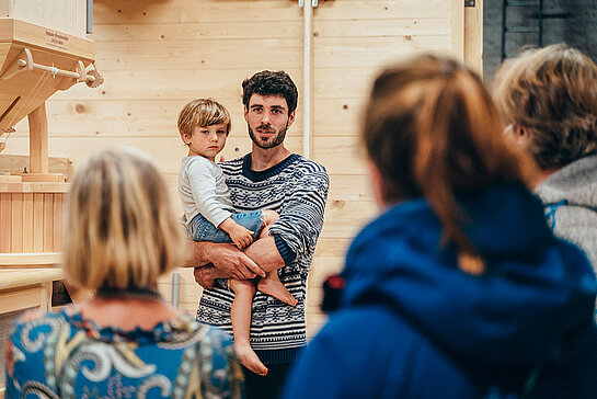 Der Landwirt Simon Reiter erklärt den Besucher*innen mit Kind auf dem Arm neben einer hölzernen Mühle die Verfahren.