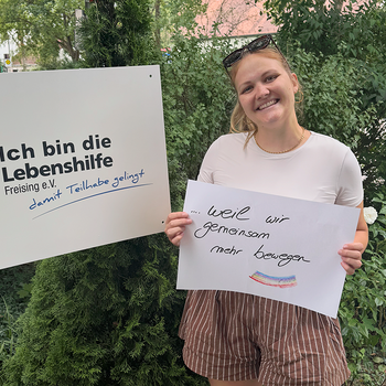 Katharina Roth feiert die Demokratie, weil wir gemeinsam mehr bewegen. Foto: Lebenshilfe Eine junge Frau mit Brille im Haar steht im Grünen und hält ein Schild, daneben hält jemand ein Schild "Ich bin die Lebenshilfe Freising e.V.