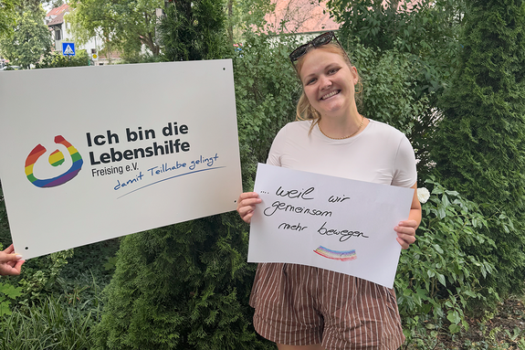 Katharina Roth feiert die Demokratie, weil wir gemeinsam mehr bewegen. Foto: Lebenshilfe Eine junge Frau mit Brille im Haar steht im Grünen und hält ein Schild, daneben hält jemand ein Schild "Ich bin die Lebenshilfe Freising e.V.