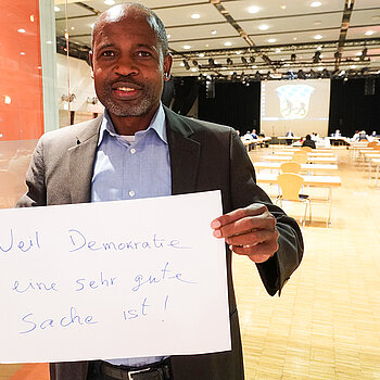 Migrationsreferent Samuel Fosso ält ein Schild mit der Aufschrift: "weil Demokrate eine sehr gute Sache ist."