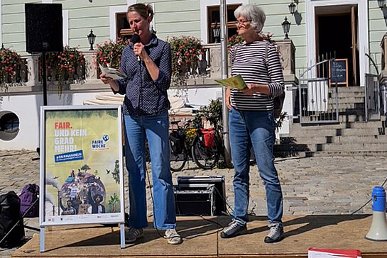 zwei Frauen auf einem Podium in der Sonne mit Mikro und einem Plakat: Fair. Und kein Grad mehr!