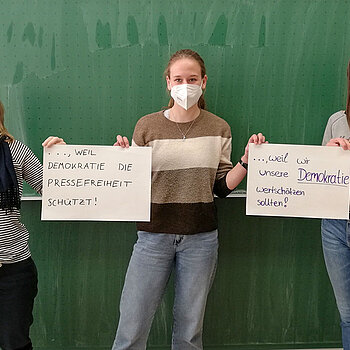 3 Schülerinnen stehen vor einer Tafel und halten 2 Schilder mit der Aufschrift. "weil Demokratie die Pressefreiheit schützt" und "weil wir unser Demokratie wertschätzen sollten"