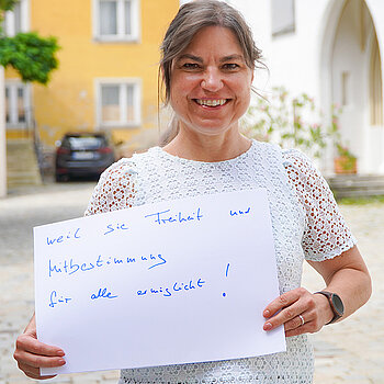 Oberbürgermeisterin Monika Schwind. Foto Stadt Freising. Eine lachende Frau mit weißem Oberteil und Schild im Freien.