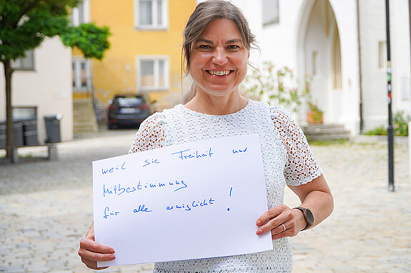 Oberbürgermeisterin Monika Schwind. Foto Stadt Freising. Eine lachende Frau mit weißem Oberteil und Schild im Freien.