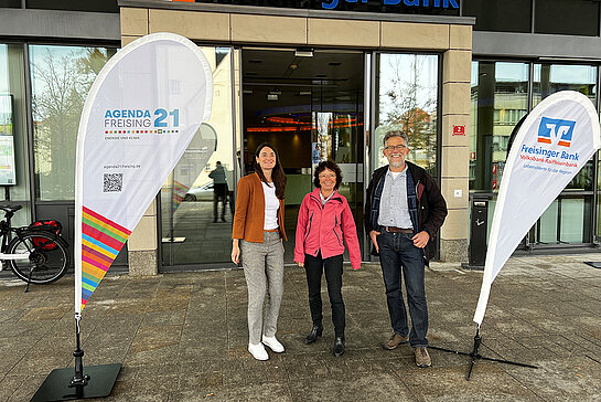 Drei Menschen vor der Freisinger Bank; eingerahmt von zwei Beachflags