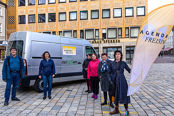 Auf den Marienplatz ein grauer Transporter mit Agenda-Logo, davor mehrere Personen mit einer Beachflag mit Agenda-Logo