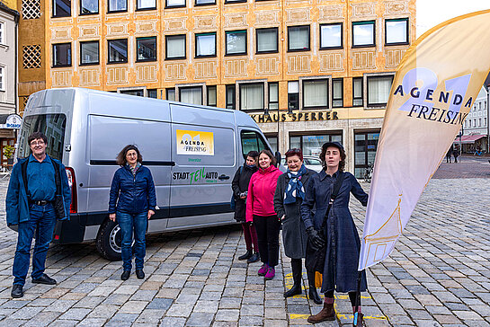 Auf den Marienplatz ein grauer Transporter mit Agenda-Logo, davor mehrere Personen mit einer Beachflag mit Agenda-Logo