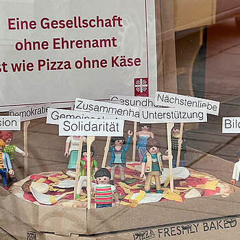 eine offenen Pizzaschachtel, darin eine Pizza und darauf Playmobil-Figuren mit Schildern: "Solidrität, Inklusion, ..."