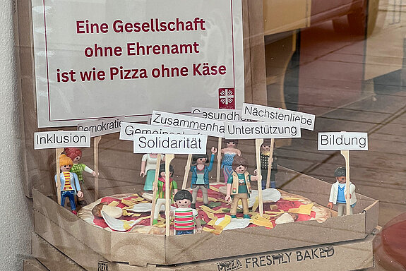 eine offenen Pizzaschachtel, darin eine Pizza und darauf Playmobil-Figuren mit Schildern: "Solidrität, Inklusion, ..."