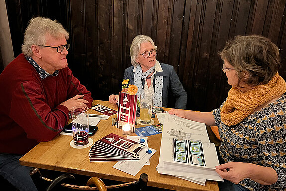 Drei Menschen sitzen an einem Tisch, vor sich Bücher und Prospekte und unterhalten sich.