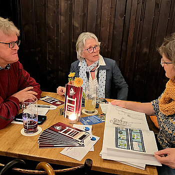 Drei Menschen sitzen an einem Tisch, vor sich Bücher und Prospekte und unterhalten sich.