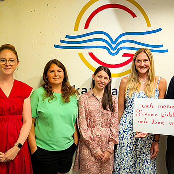 Wir feiern die Demokratie, weil meine Stimme zählt - und deine auch. Foto: D. Lepkova vor einer hellen Wand mit dem Logo des OMG (ein gelber Kreis, darin ein rotes G und ein stilisierte blaues aufgeschlagenes Buch stehen 5 Frauen und ein Mann mit Bart der ein Schild hält