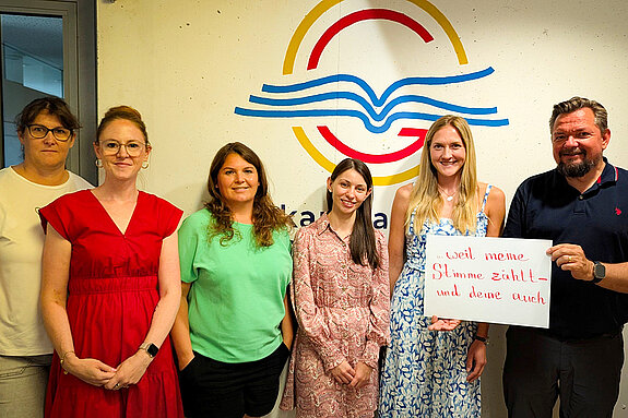 Wir feiern die Demokratie, weil meine Stimme zählt - und deine auch. Foto: D. Lepkova vor einer hellen Wand mit dem Logo des OMG (ein gelber Kreis, darin ein rotes G und ein stilisierte blaues aufgeschlagenes Buch stehen 5 Frauen und ein Mann mit Bart der ein Schild hält