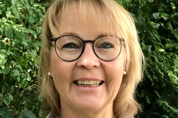 eine lachende blonde Frau mit Brille vor Büschen