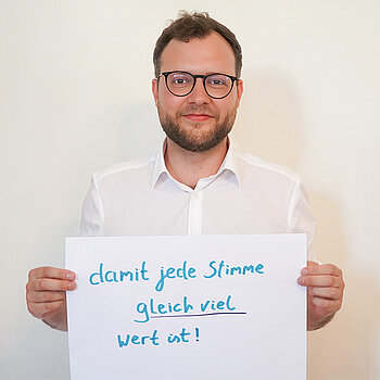 Zweiter Bürgermeister Nico Heitz. Foto: Stadt Freising Ein Mann in einem weißen Hemd zeigt ein Schild mit der Aufschrift "damit jede Stimme gleich viel wert ist!"