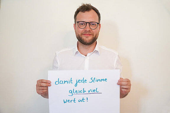 Zweiter Bürgermeister Nico Heitz. Foto: Stadt Freising Ein Mann in einem weißen Hemd zeigt ein Schild mit der Aufschrift "damit jede Stimme gleich viel wert ist!"