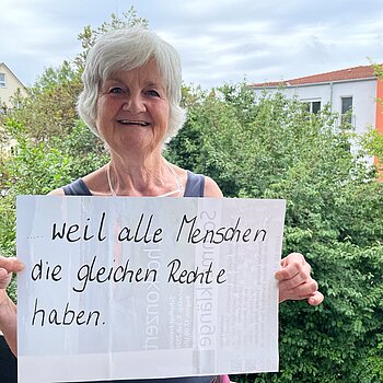 Monika Hobmair (Agenda21) feiert die Demokratie, weil alle Menschen die gleichen Rechte haben. Foto: privat Eine Frau mit kurzen grauen Haaren steht auf einem Balkon, Bäume im Hintergrund und hält ein Schild.