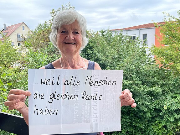 Monika Hobmair (Agenda21) feiert die Demokratie, weil alle Menschen die gleichen Rechte haben. Foto: privat Eine Frau mit kurzen grauen Haaren steht auf einem Balkon, Bäume im Hintergrund und hält ein Schild.