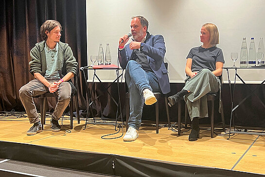 zwei Männer und eine Frau sitzen auf Stühlen nebeneinander und reden miteinander