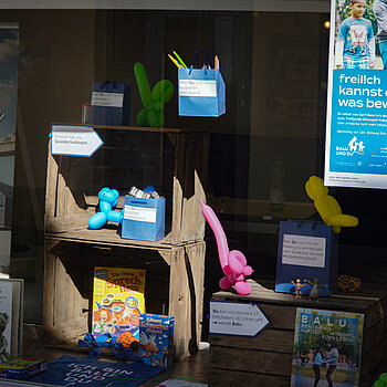 Blau Papiertüten mit Spielmaterial, Stiften und Plätzchenformen darin auf Kisten und mit Luftballons
