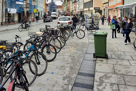 Untere Hauptstraße mit vielen Rädern in einer Reihe abgestellt, einer grünen Tonne auf der Sehbehindertenleitlinie und weiter hinten einen Transportwagen mit gelben Säcken