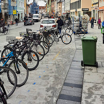 Untere Hauptstraße mit vielen Rädern in einer Reihe abgestellt, einer grünen Tonne auf der Sehbehindertenleitlinie und weiter hinten einen Transportwagen mit gelben Säcken