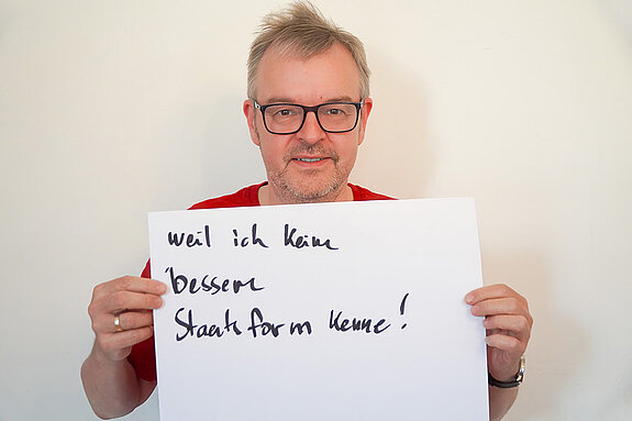 Stadtratsmitglied Ulrich Vogl. Foto: Stadt Freising Ein Mann mit Brille zeigt ein Schild mit der Aufschrift "weil ich keine bessere Staatsform kenne!"