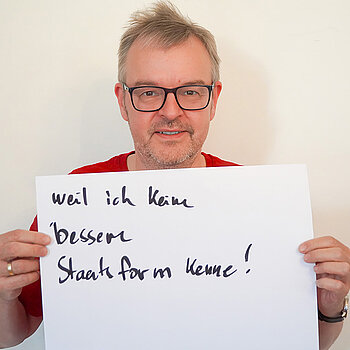 Stadtratsmitglied Ulrich Vogl. Foto: Stadt Freising Ein Mann mit Brille zeigt ein Schild mit der Aufschrift "weil ich keine bessere Staatsform kenne!"