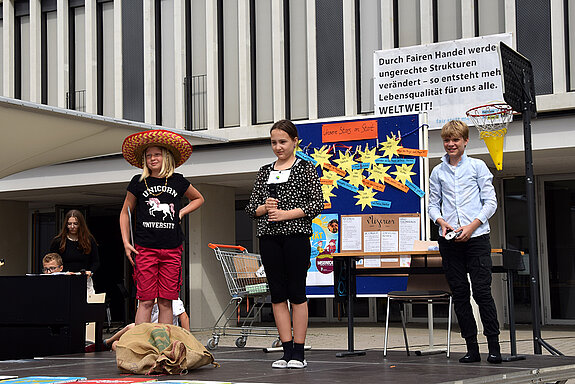 3 Kinder stehen auf einer Bühne, hinter ihnen ein Plakat, eine Pinnwand und ein Einkaufswagen
