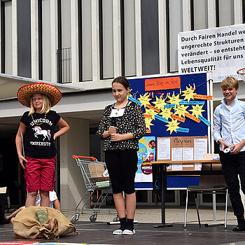 3 Kinder stehen auf einer Bühne, hinter ihnen ein Plakat, eine Pinnwand und ein Einkaufswagen
