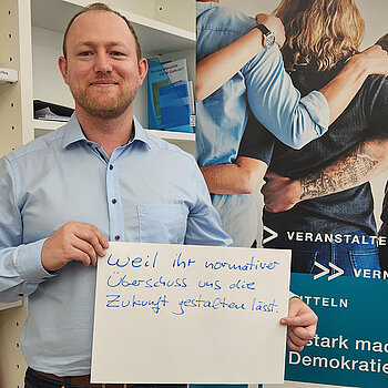 Kai Kallbach feiert die Demokratie, weil ihr normativer Überschuss uns die Zukunft gestalten lässt. Foto: Domberg Akademie Ein Mann im hellblauen Hemt steht neben einer Bücherwand und hält ein Schild.