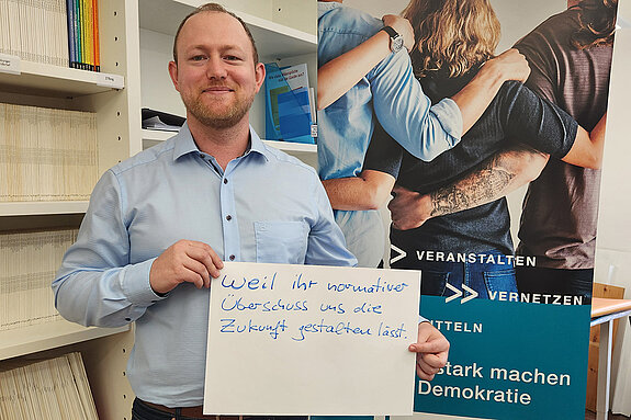 Kai Kallbach feiert die Demokratie, weil ihr normativer Überschuss uns die Zukunft gestalten lässt. Foto: Domberg Akademie Ein Mann im hellblauen Hemt steht neben einer Bücherwand und hält ein Schild.