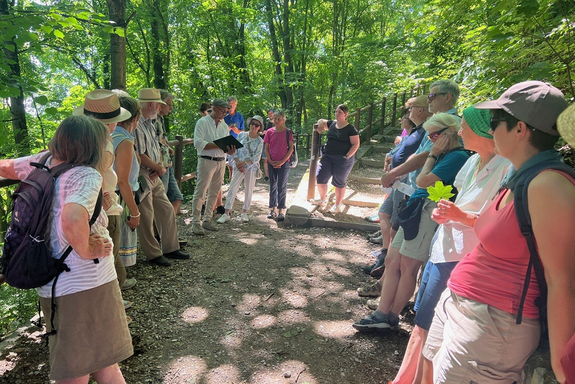 eine Gruppe Menschen in sommerlicher Kleidung steht auf zwei Seiten eines Weges unter einem Blätterdach. Ein Mann hat ein Buch in der Hand und liest daraus vor.